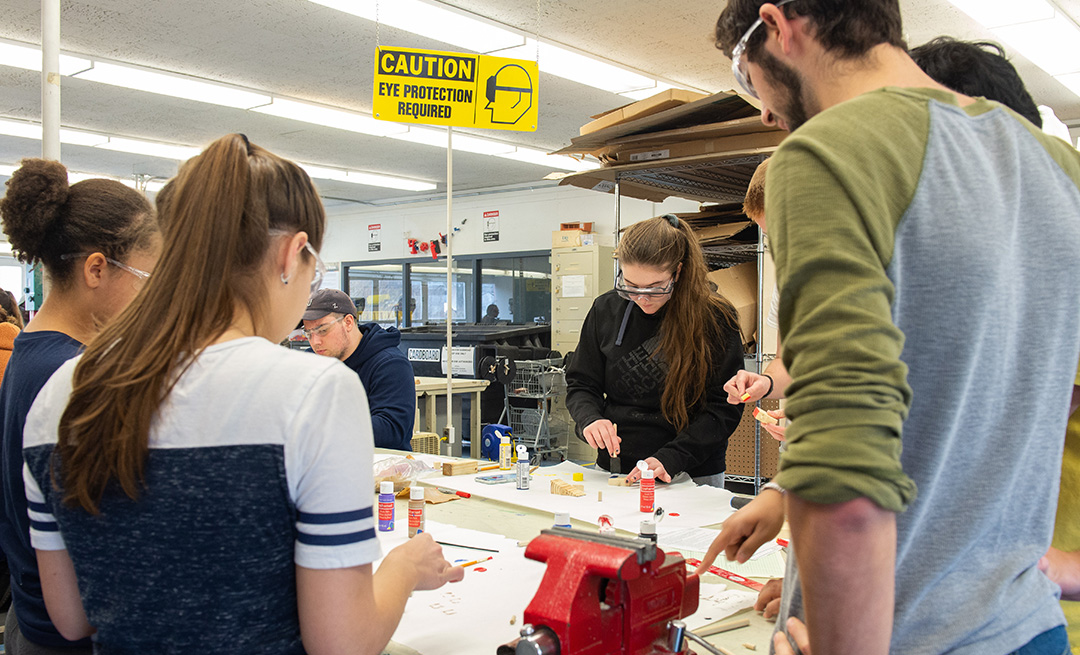 Students stand around a work bench, working with glue and schematics