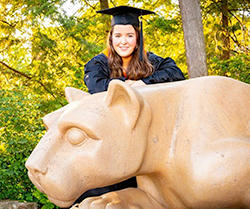 A woman wearing a blue graduation cap and gown poses for a photo on the Penn State Nittany Lion, a sculpture of a mountain lion. 