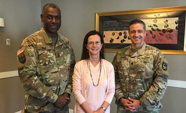 Two men in Army uniforms and a woman in a pink sweater stand in front of a grey and white wall