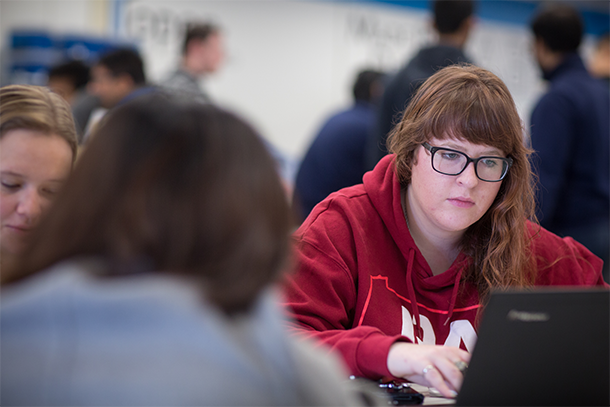 Woman in red sweatshirt sits at a table and looks at a computer.