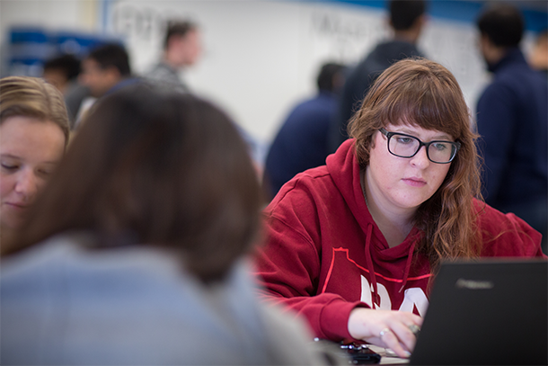 Woman in red sweatshirt sits at a table and looks at a computer. 