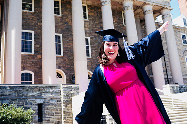 White woman in pink dress and blue graduation cap and gown smiles in front of a brick building
